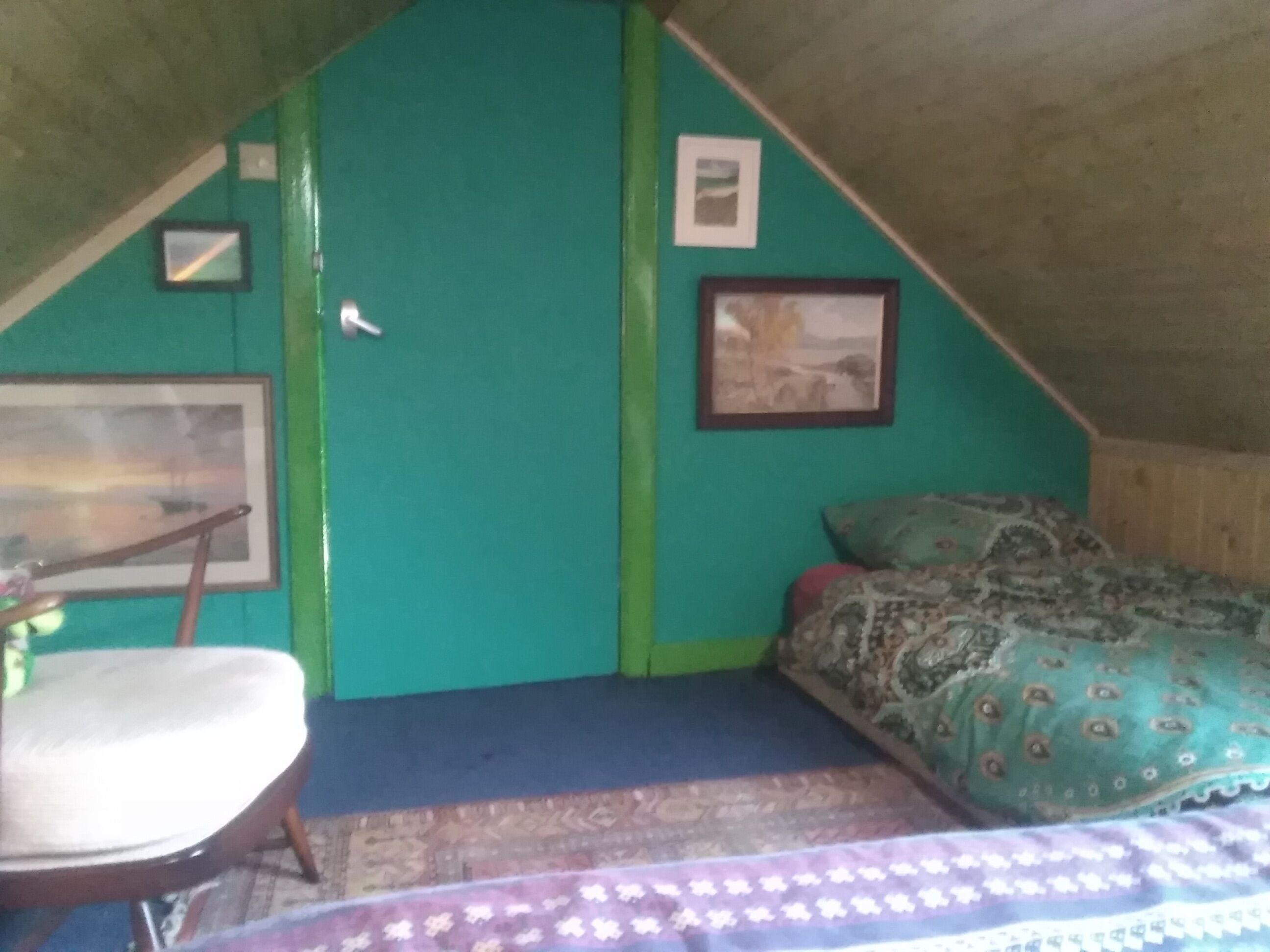 A small, cozy room with a sloped ceiling, a single bed with patterned bedding, a chair, and a framed picture on the wall.