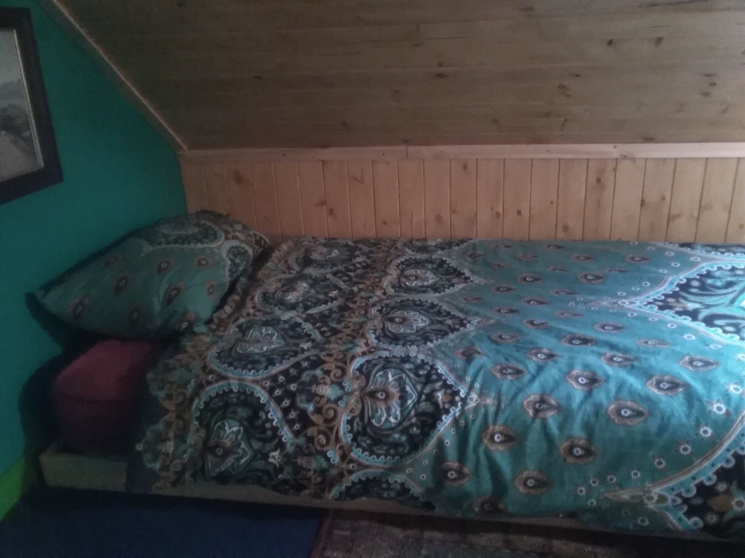 A small, cozy room with a wooden ceiling and walls, a bed with patterned bedding, and a single red cushion.