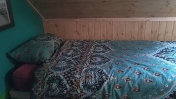A small, cozy room with a wooden ceiling and walls, a bed with patterned bedding, and a single red cushion.