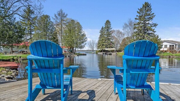 Two blue Adirondack chairs on a wooden dock overlooking a calm lake with trees and a house in the background.