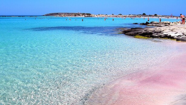 Una playa con agua turquesa cristalina, costa rocosa y gente nadando.