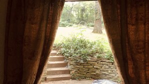 Blick durch ein Fenster mit Vorhängen, der eine Steintreppe zu einer Grünanlage mit Pflanzen und einem überdachten Gegenstand im Erdgeschoss zeigt.