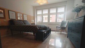 Ein Schlafzimmer mit Bett, Sessel, Kommode und Fenster mit Blick auf Gebäude.