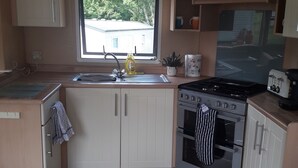 A compact kitchen with a window, a sink, a stove, and wooden cabinets. - 3-bedroom Caravan at Thorness bay (Cowes)