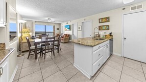 A spacious kitchen with a granite countertop, white cabinets, and a large island. There is a dining area with a table and chairs, and a view of the ocean through large windows. - Harbour Place 313 (Orange Beach)