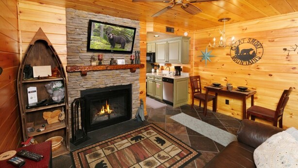 A cozy cabin interior with a stone fireplace, wooden walls, a television mounted above it, a dining area with a table and chairs, and a kitchen area with cabinets and a microwave.