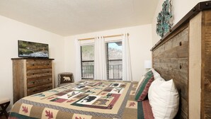 A bedroom with a wooden headboard, a quilted bedspread, a wooden dresser, a television on top, and a window with curtains. - Oak Square 305 - Ask About Free Attraction Tickets! (Gatlinburg)