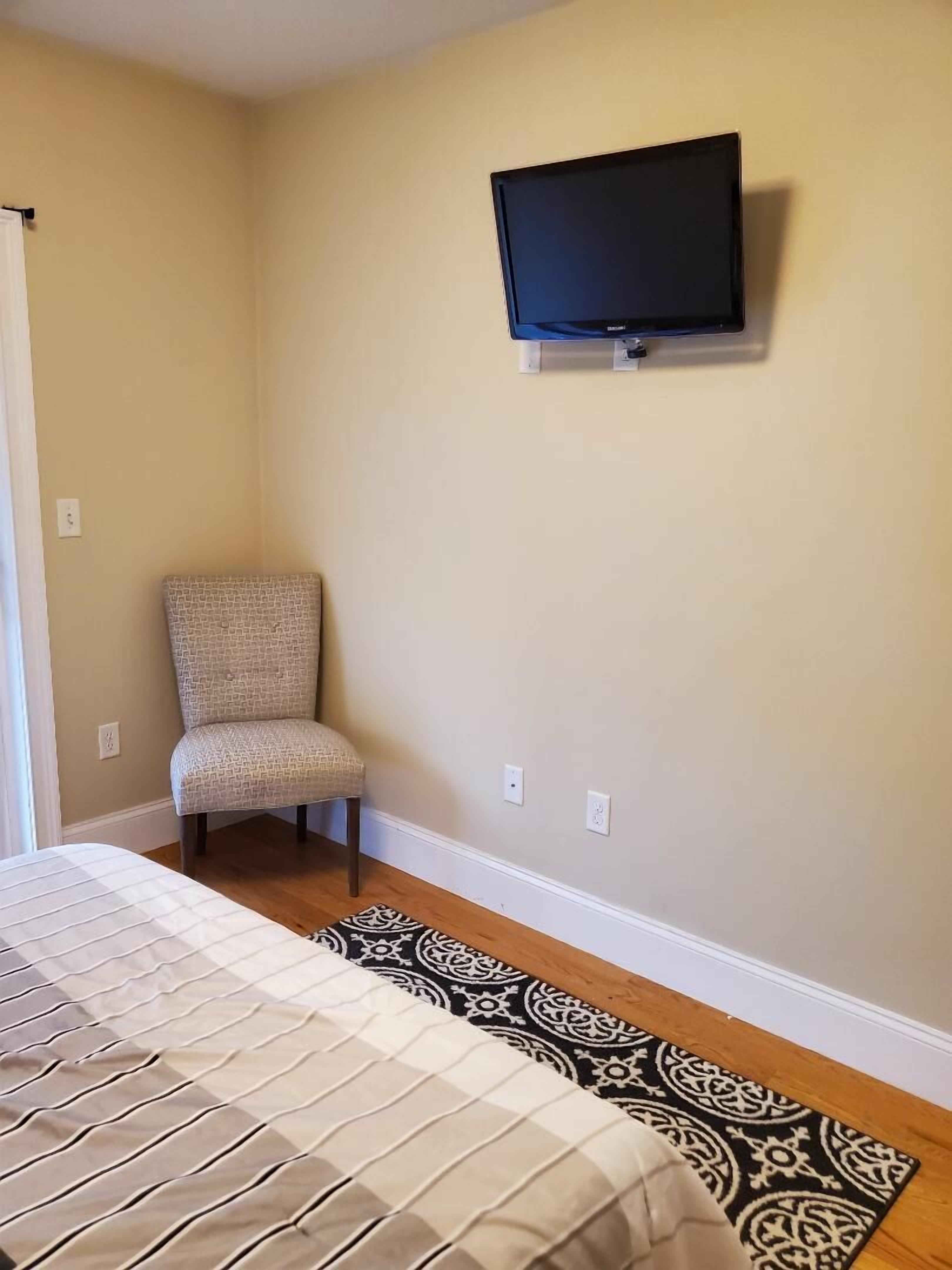 A bedroom with a bed, a chair, a television mounted on the wall, and a patterned rug on the floor.