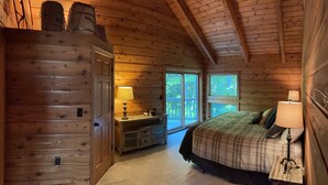 Un dormitorio tipo cabaña de madera con una cama, dos lámparas de noche, un aparador y una ventana con vista a los árboles.