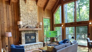 Una sala de estar acogedora con chimenea de piedra, piso de madera y ventanas amplias con vistas al bosque.