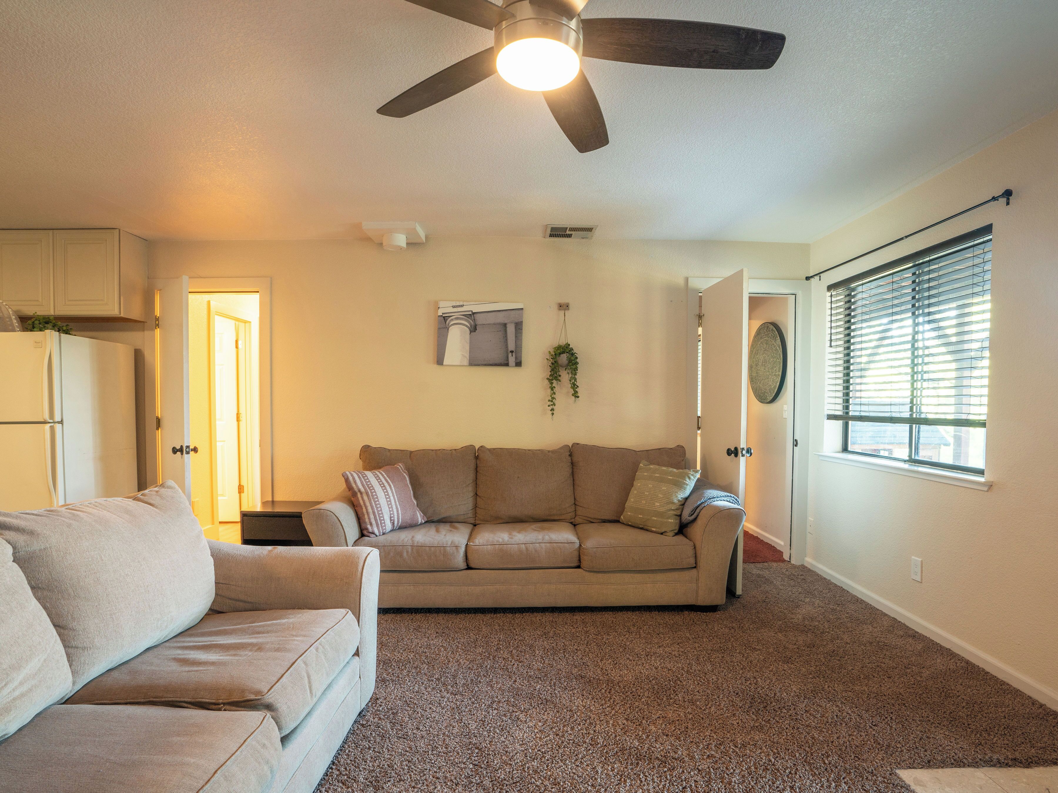 A living room with a beige sofa, a light-colored couch, a ceiling fan, and a window with blinds.