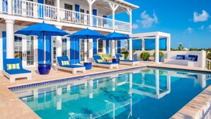 A poolside area with lounge chairs and umbrellas, a clear blue pool, and a white building with balconies. - Beautiful Home by Taylor Bay Beach (Providenciales)