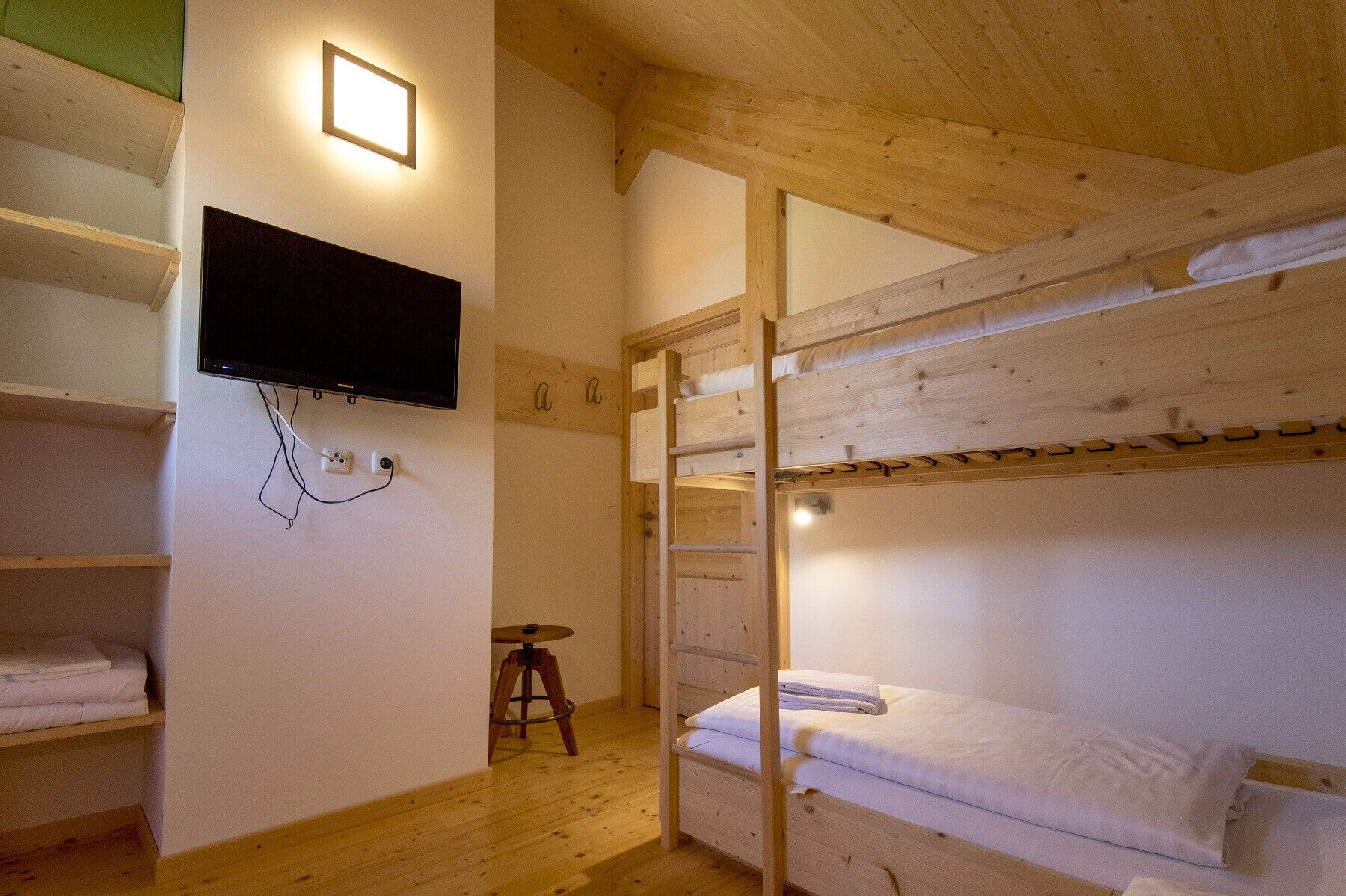 Habitación con literas, un televisor montado en la pared, vigas de madera y un taburete pequeño.