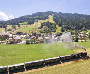Un train à vapeur traverse une zone rurale, avec des maisons et une colline boisée en arrière-plan.
