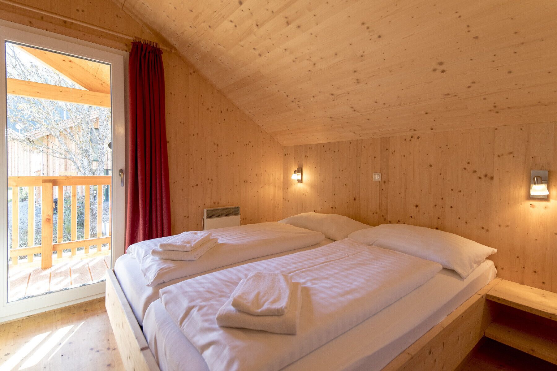 A wooden cabin room with two beds, a window with a red curtain, and a wooden deck outside.