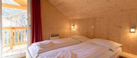 A wooden cabin room with two beds, a window with a red curtain, and a wooden deck outside.