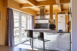 Une cuisine moderne avec des poutres en bois, un îlot central doté de tabourets de bar et une porte-fenêtre coulissante donnant sur un espace extérieur.