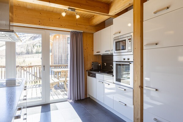 Una cocina moderna con armarios blancos, horno empotrado y zona de comedor con terraza de madera.