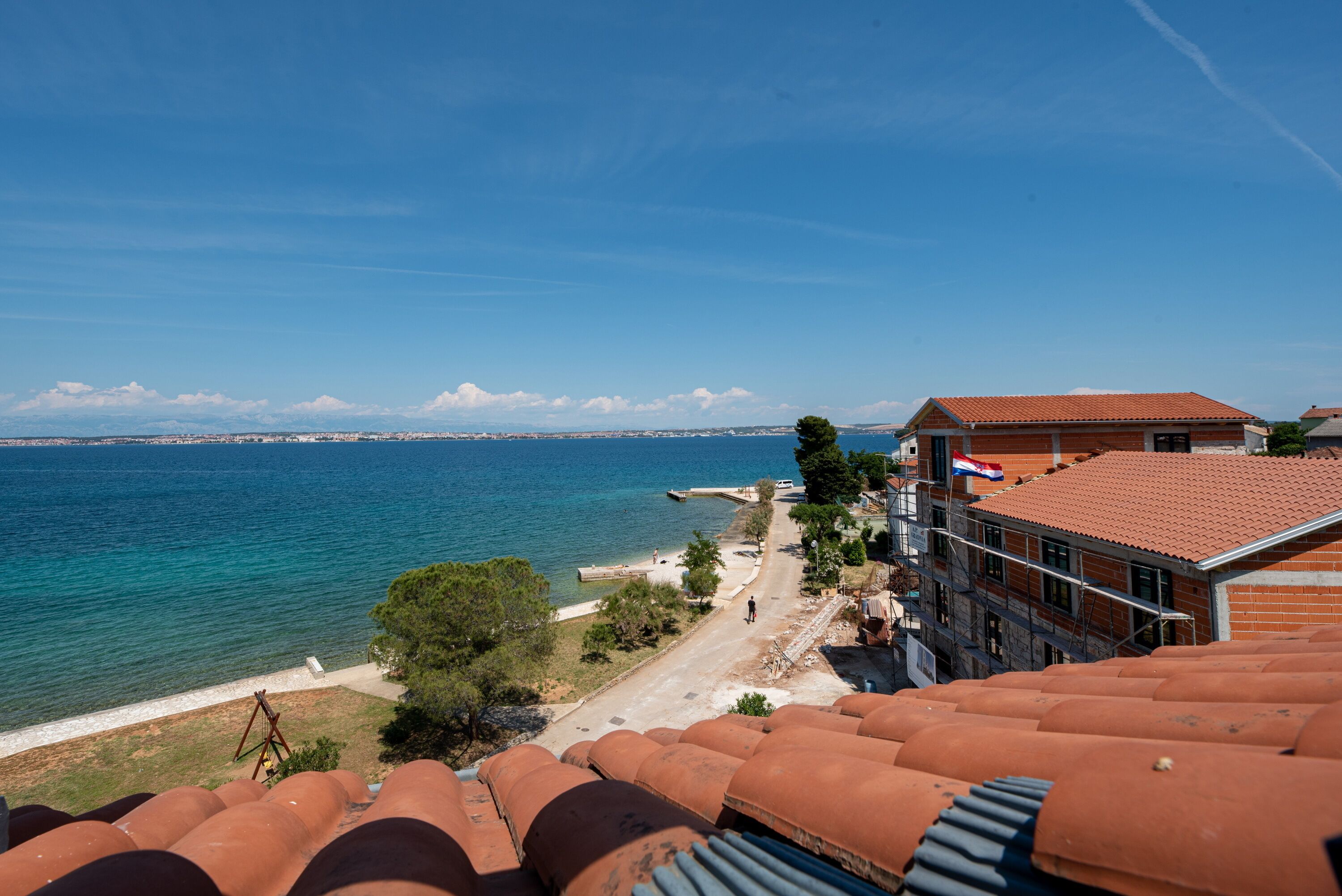 Paesaggio costiero con edifici dalle tegole color terracotta, un mare blu limpido e una costa in lontananza.