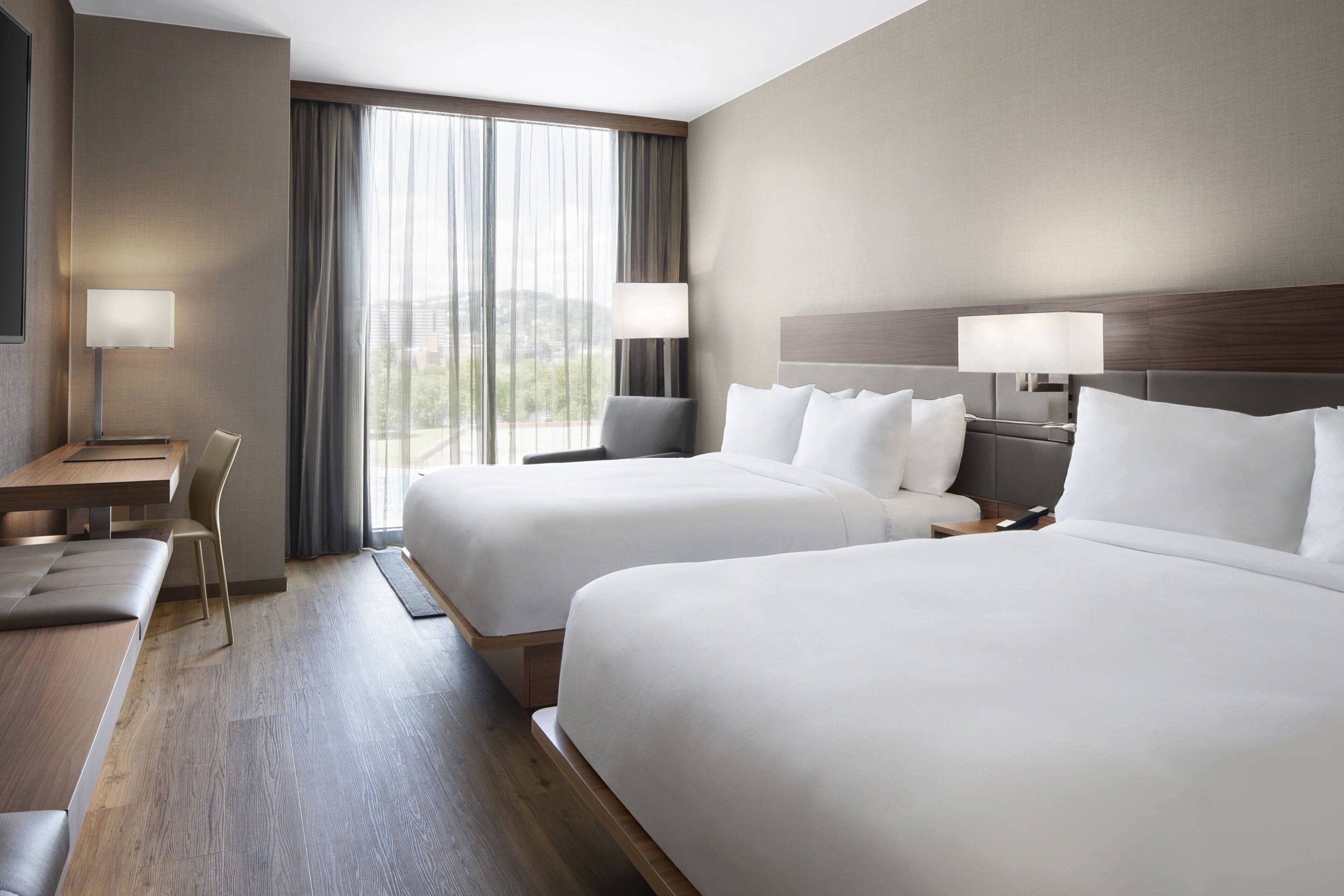 A hotel room with two beds, a desk, a chair, and a large window with curtains.