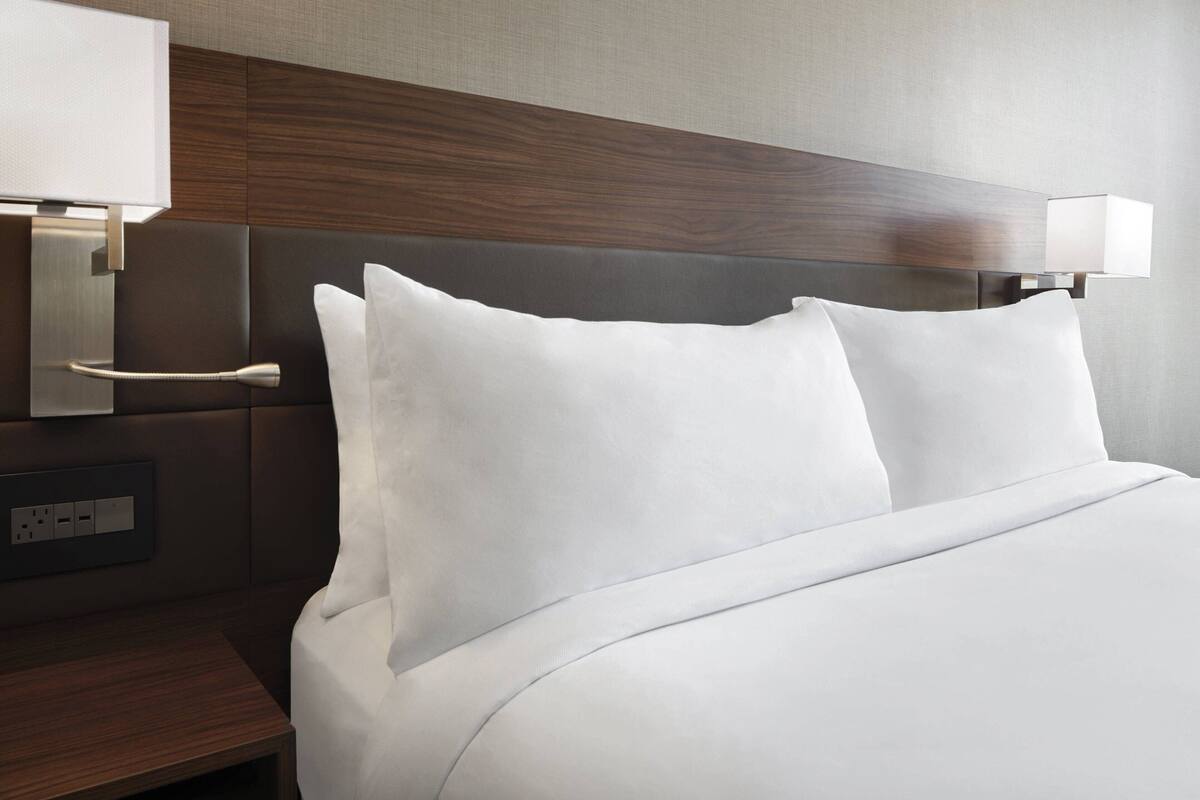 Een hotelkamer met een net opgemaakt bed, een nachtkastje met een lamp en een wandcontactdoos.