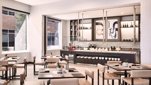 A modern dining area with tables and chairs, a kitchen counter with various items, and a large window providing natural light.