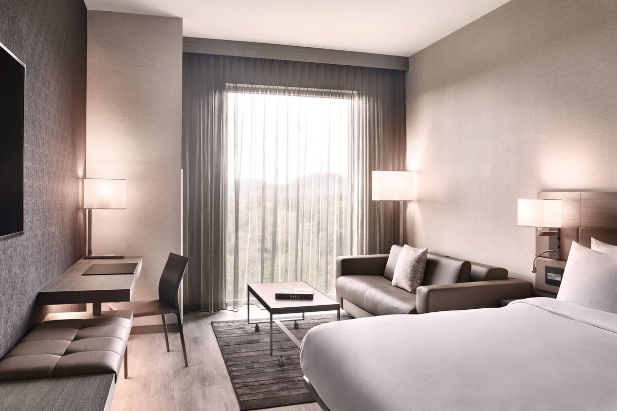Una habitación de hotel moderna con una cama grande, un sofá, una mesita y un escritorio. Hay dos lámparas a cada lado de la habitación.