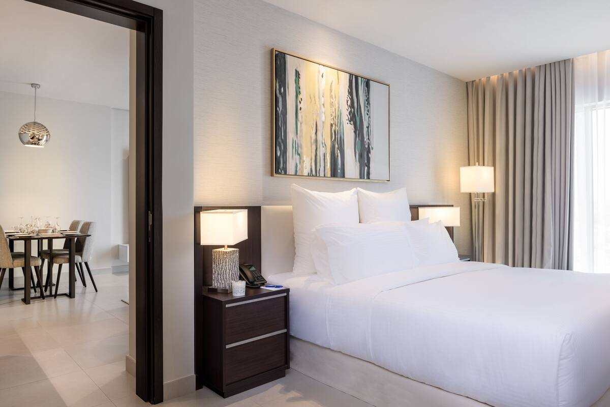 Habitación de hotel con cama, dos lámparas de noche, una mesita de noche, un cuadro en la pared y vista a un comedor.