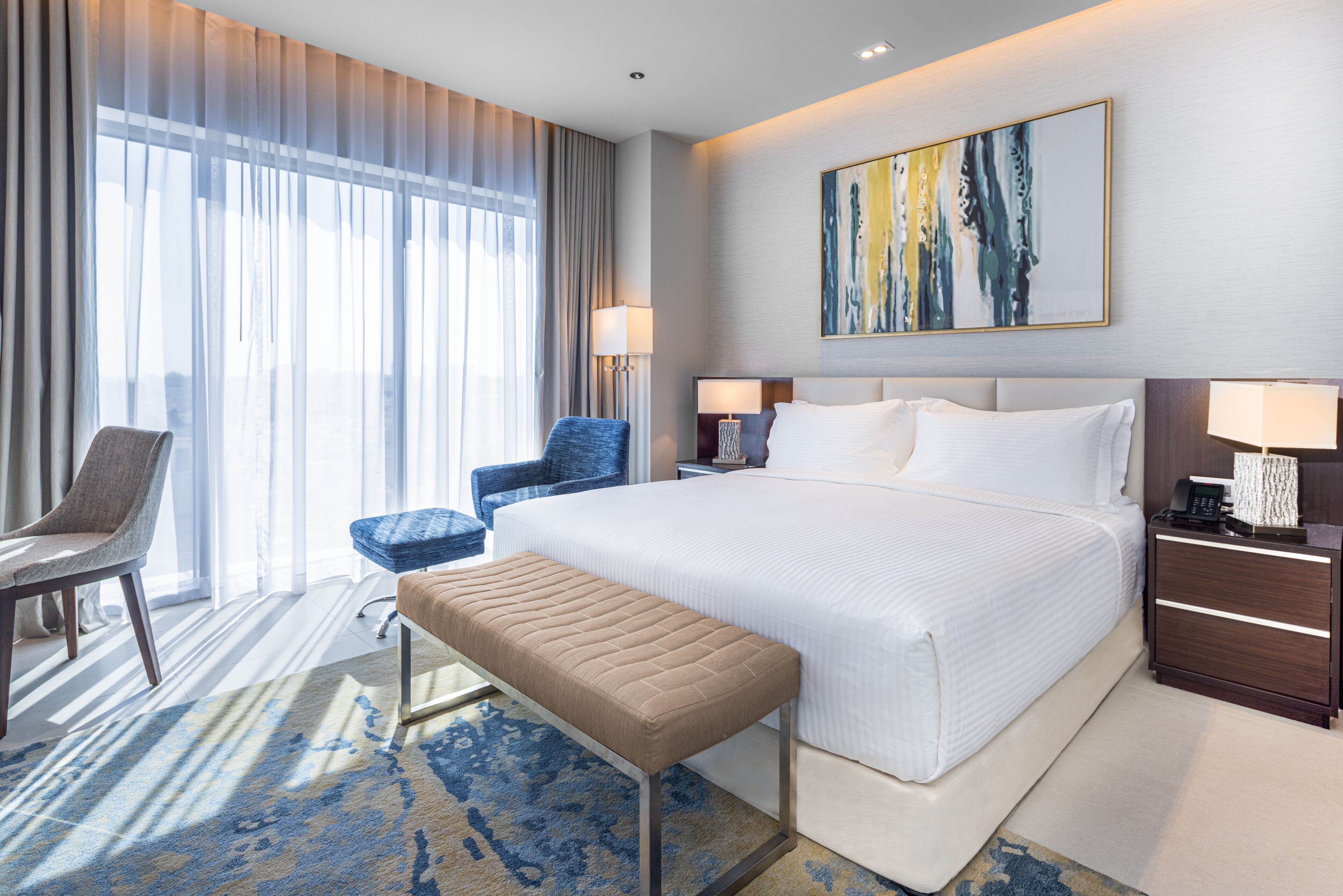 A hotel room with a large bed, a bench, a chair, a desk, and a painting on the wall.