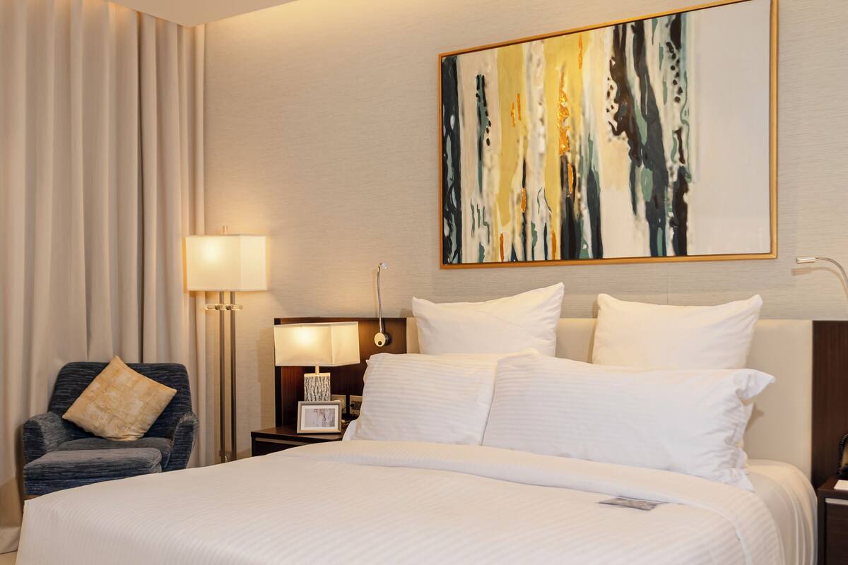 Ein Hotelzimmer mit einem großen Bett, einem Sessel, einem Nachttisch, einer Lampe und einem Gemälde an der Wand.