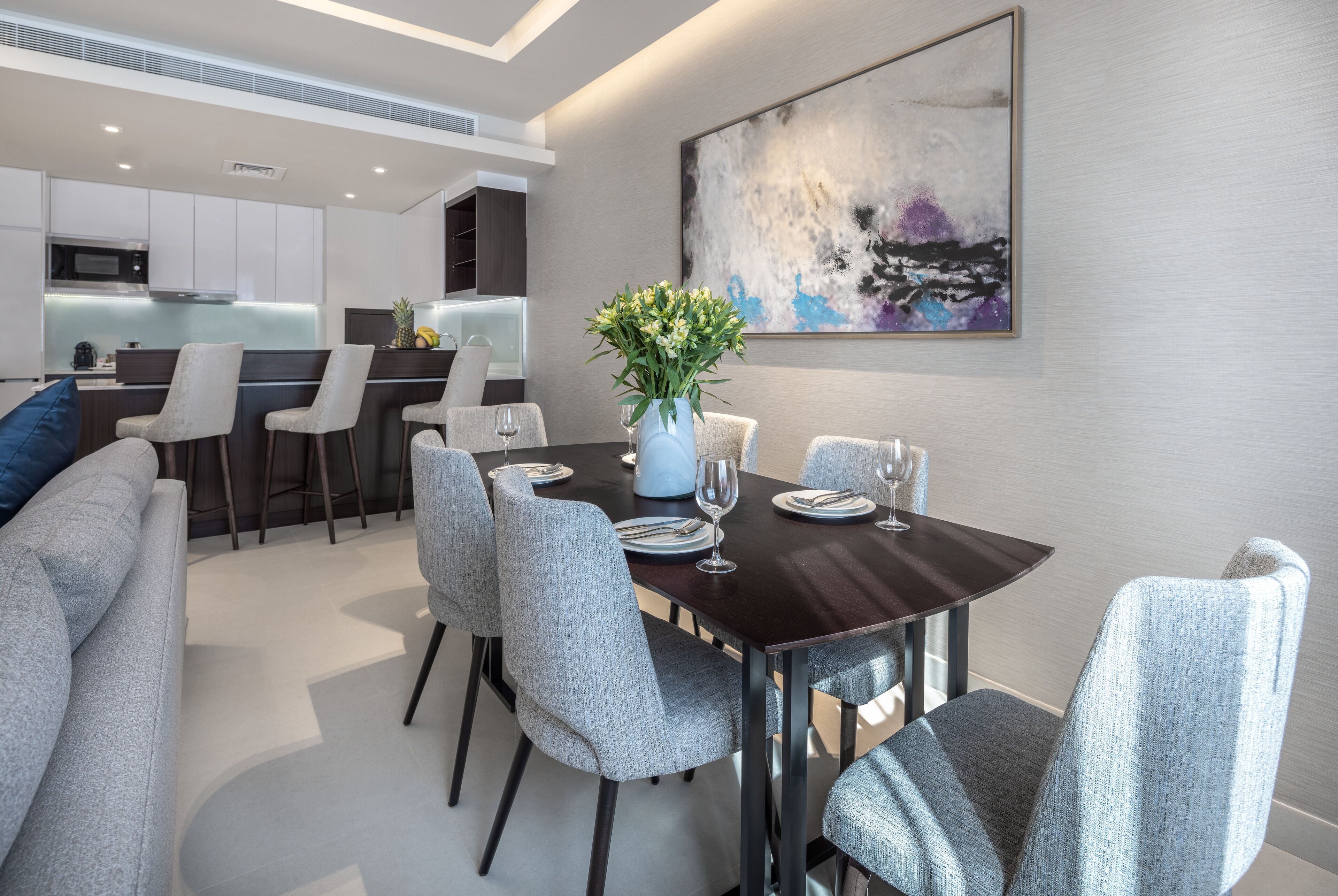 Un comedor moderno con una mesa de madera oscura, sillas tapizadas de color gris y un jarrón de flores. Se ve una cocina al fondo con armarios blancos y un microondas.