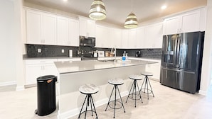 A modern kitchen with white cabinets, a black refrigerator, a large island with stools, and pendant lighting. - Luxury 6 bed South-Facing Pool, Game Room, 4 Miles to Disney World (Kissimmee)