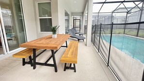 A covered patio with a wooden table and benches, a pool with a safety fence, and large windows allowing natural light. - Luxury 6 bed South-Facing Pool, Game Room, 4 Miles to Disney World (Kissimmee)