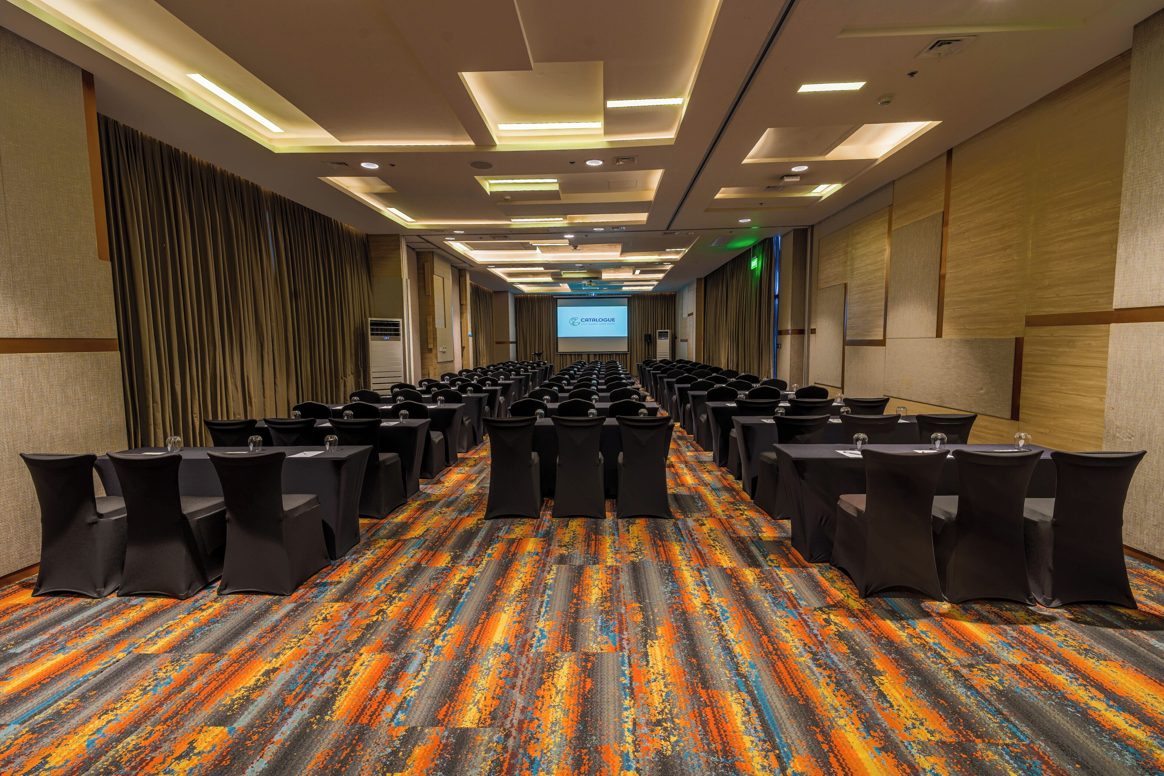 A conference room with rows of tables and chairs, a projector screen, and a colorful carpet.