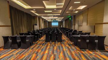 A conference room with rows of tables and chairs, a projector screen, and a colorful carpet.
