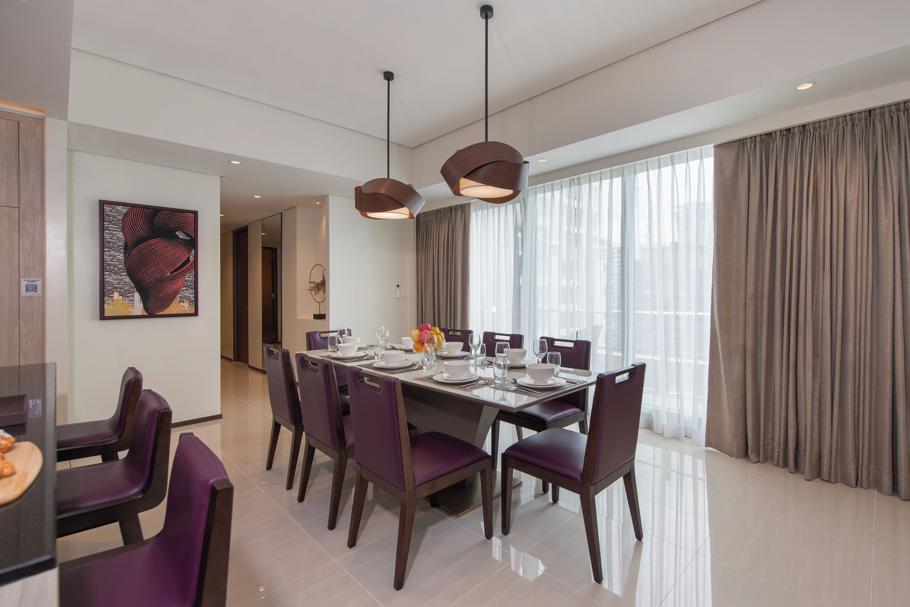 Un comedor moderno con una mesa larga dispuesta para seis personas, rodeado de sillas moradas y ventanas grandes con cortinas.