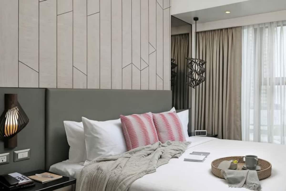 A modern hotel room with a bed, bedside table, lamp, and a view of the city through large windows.