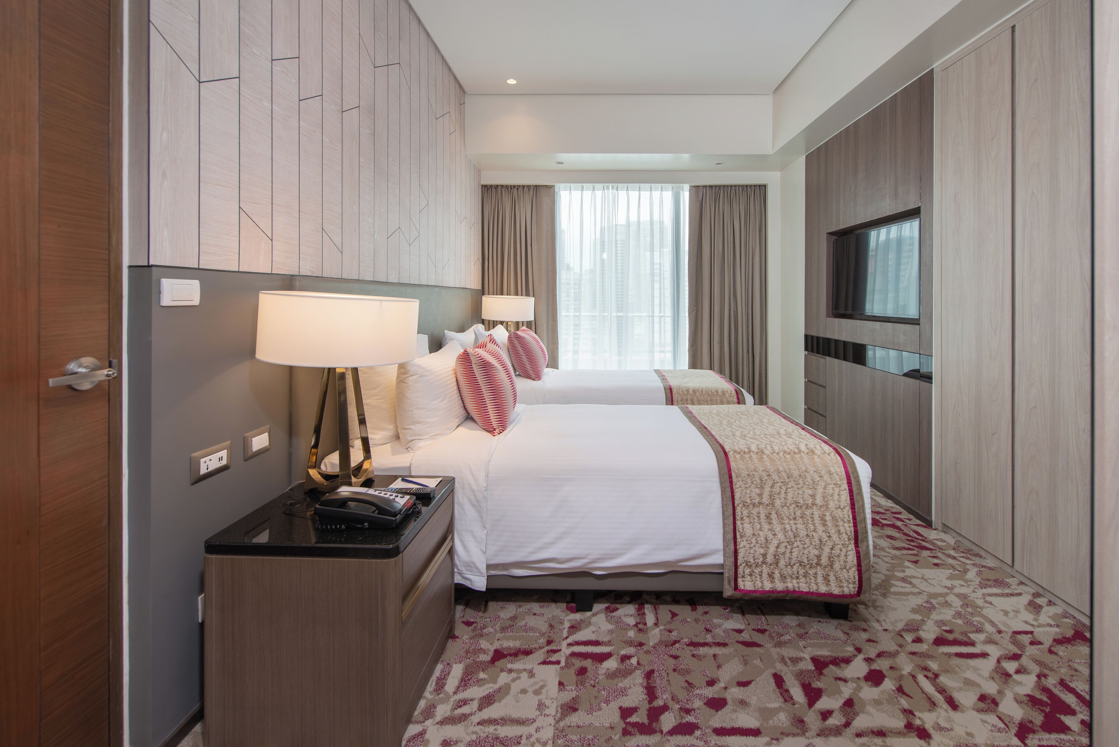 A hotel room with two beds, a desk with a lamp, a TV, and a large window with curtains.