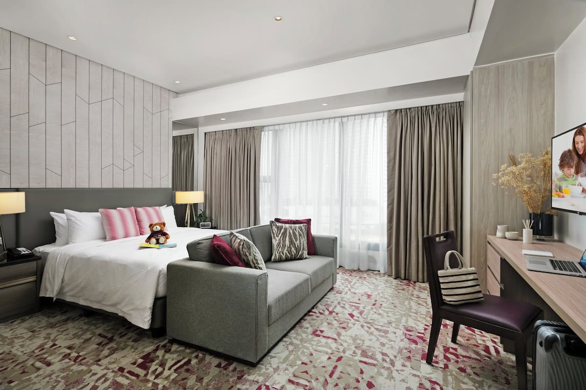 Ein modernes Hotelzimmer mit einem großen Bett, einem grauen Sofa, einem Schreibtisch mit einem Laptop und einem Stuhl.