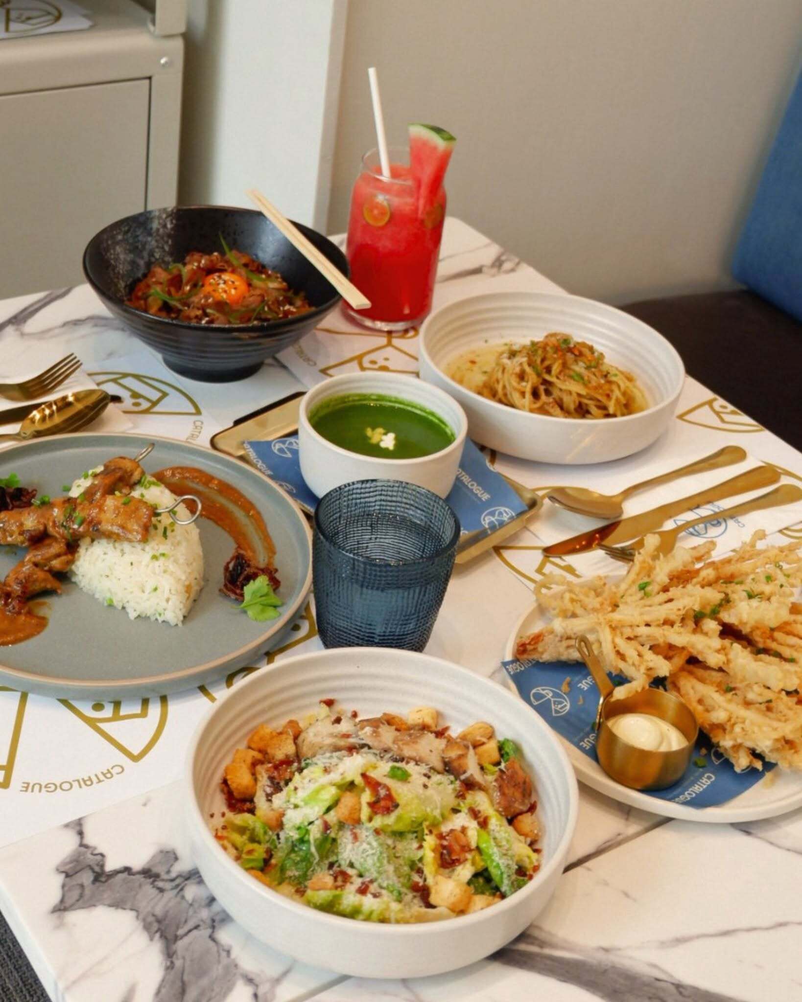 Une table garnie de différents plats, notamment du riz, des pâtes et une salade, ainsi qu’une boisson et un bol de soupe.
