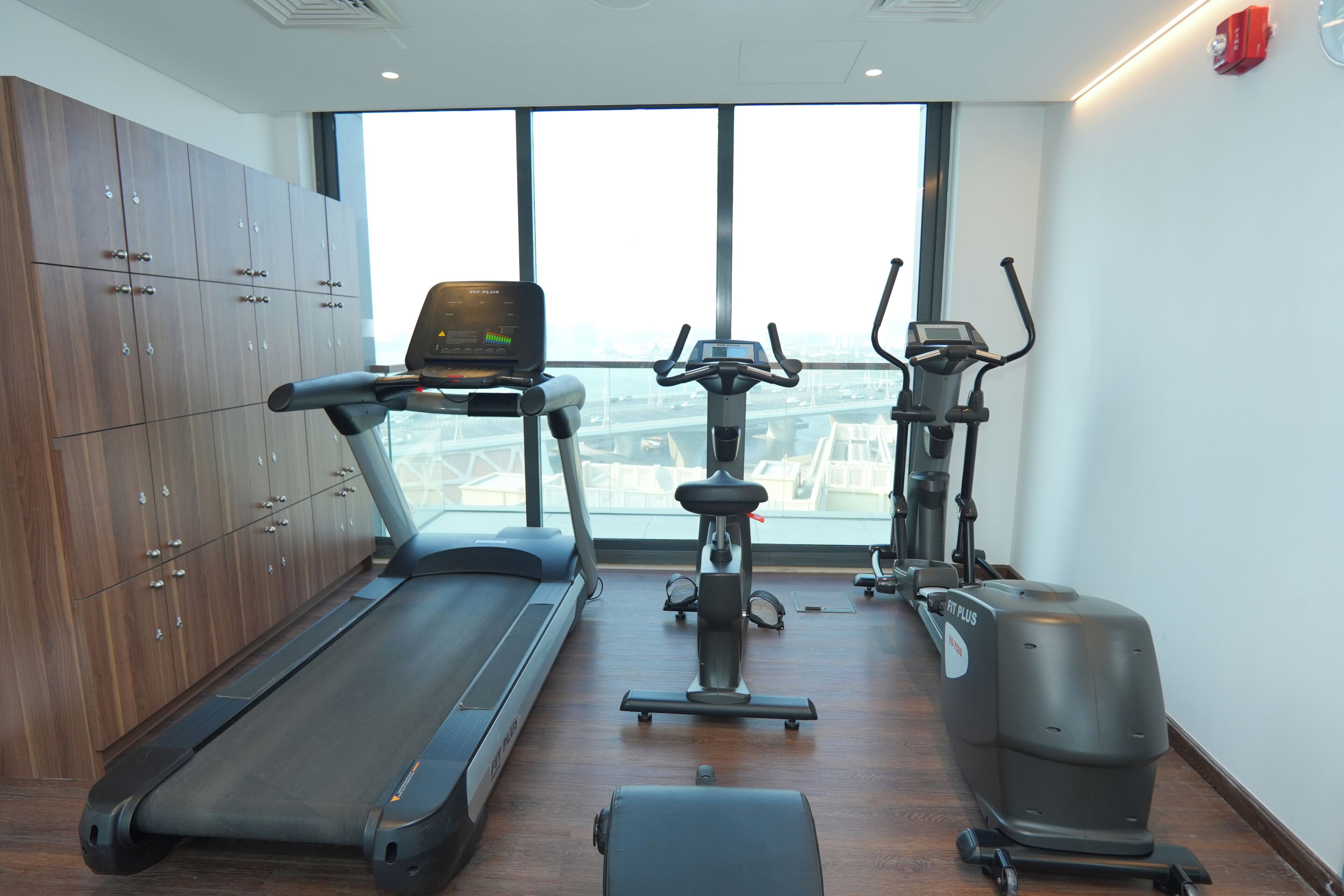 A gym with a treadmill, stationary bike, and elliptical machine, large windows, and wooden lockers.