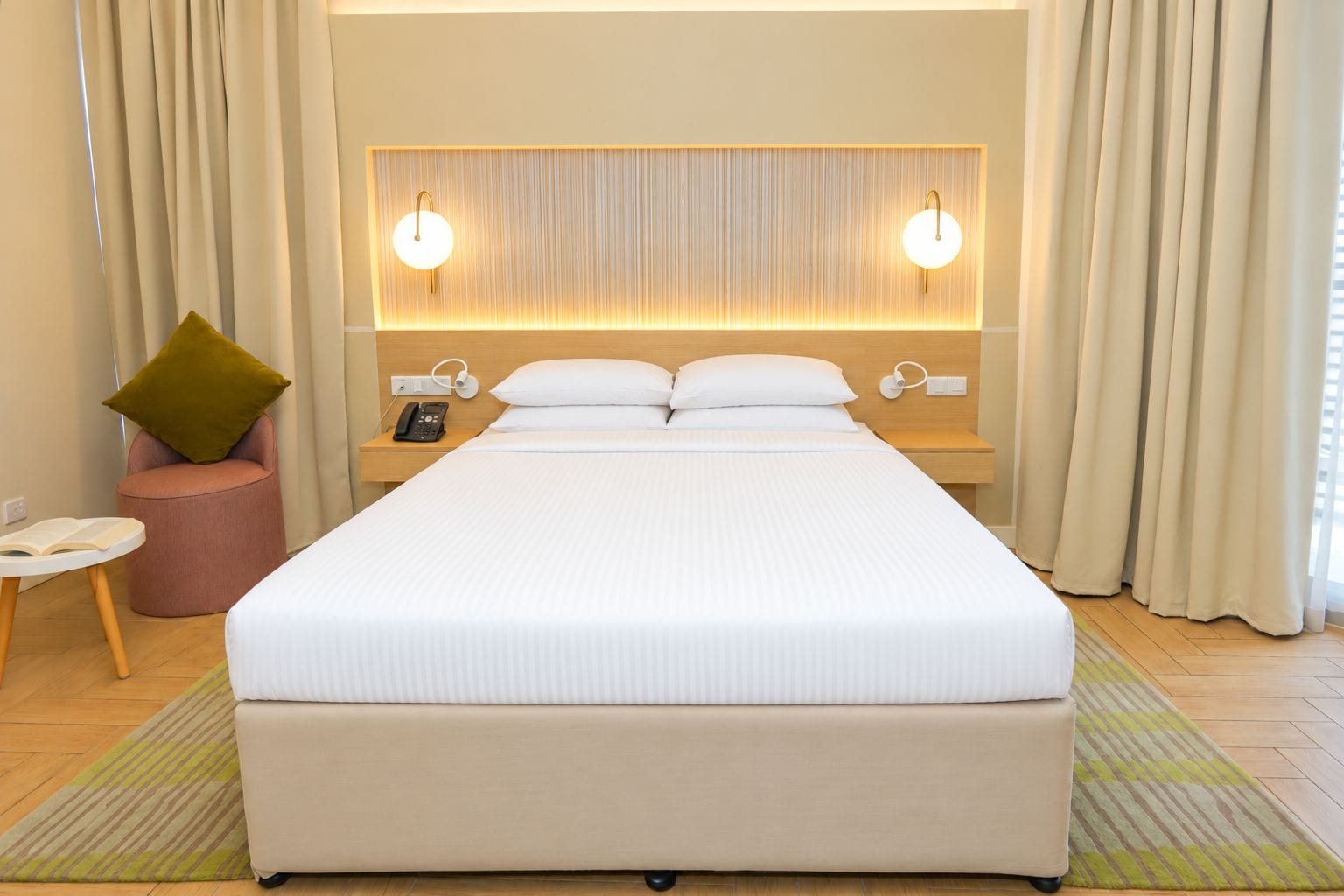 A hotel room with a large bed, two bedside lamps, a bedside phone, a small table with a book, and a green pillow.