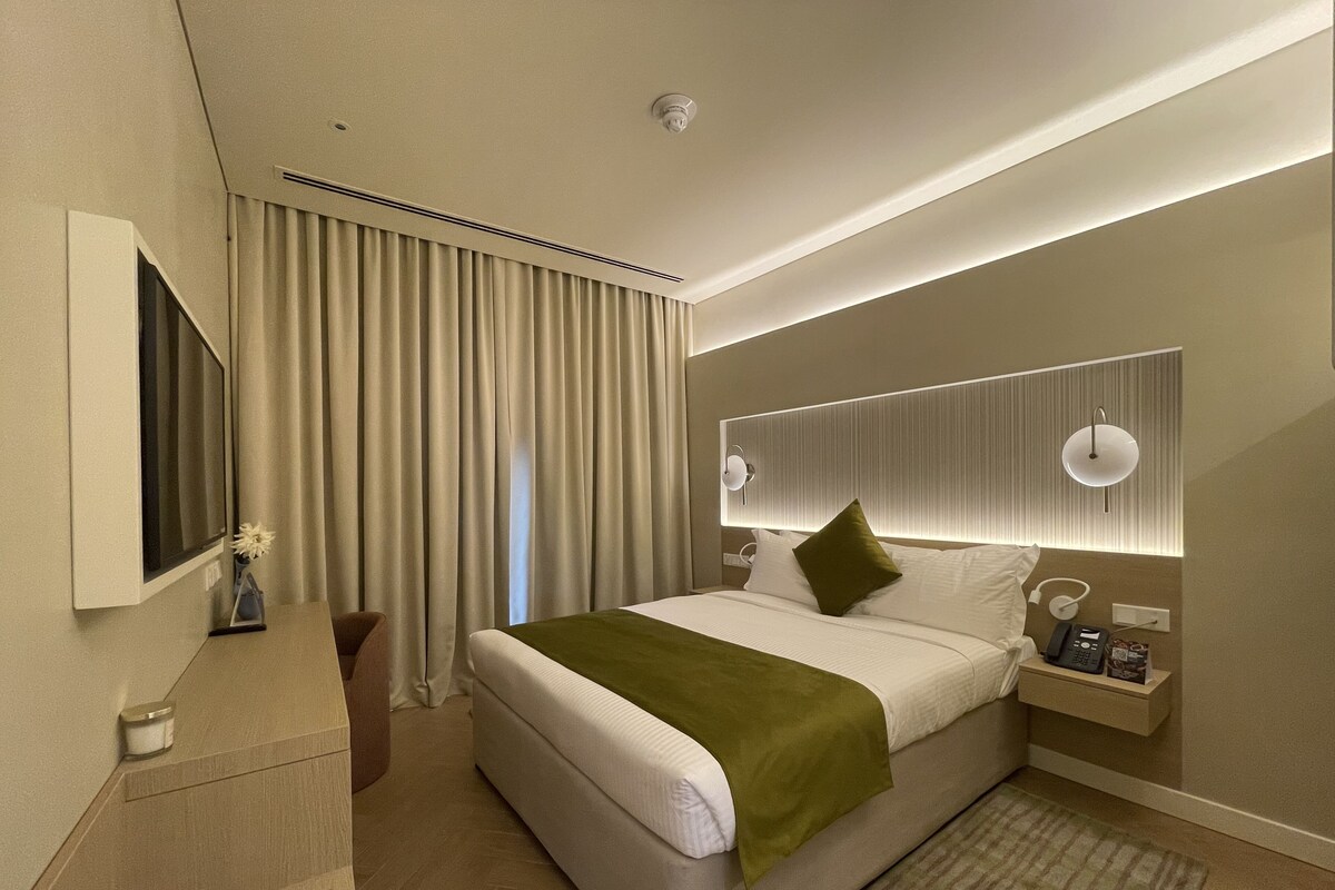 Ein modernes Hotelzimmer mit einem großen Bett, einem Nachttisch mit Lampe, einem Flachbildfernseher und einem großen Fenster mit Vorhängen.