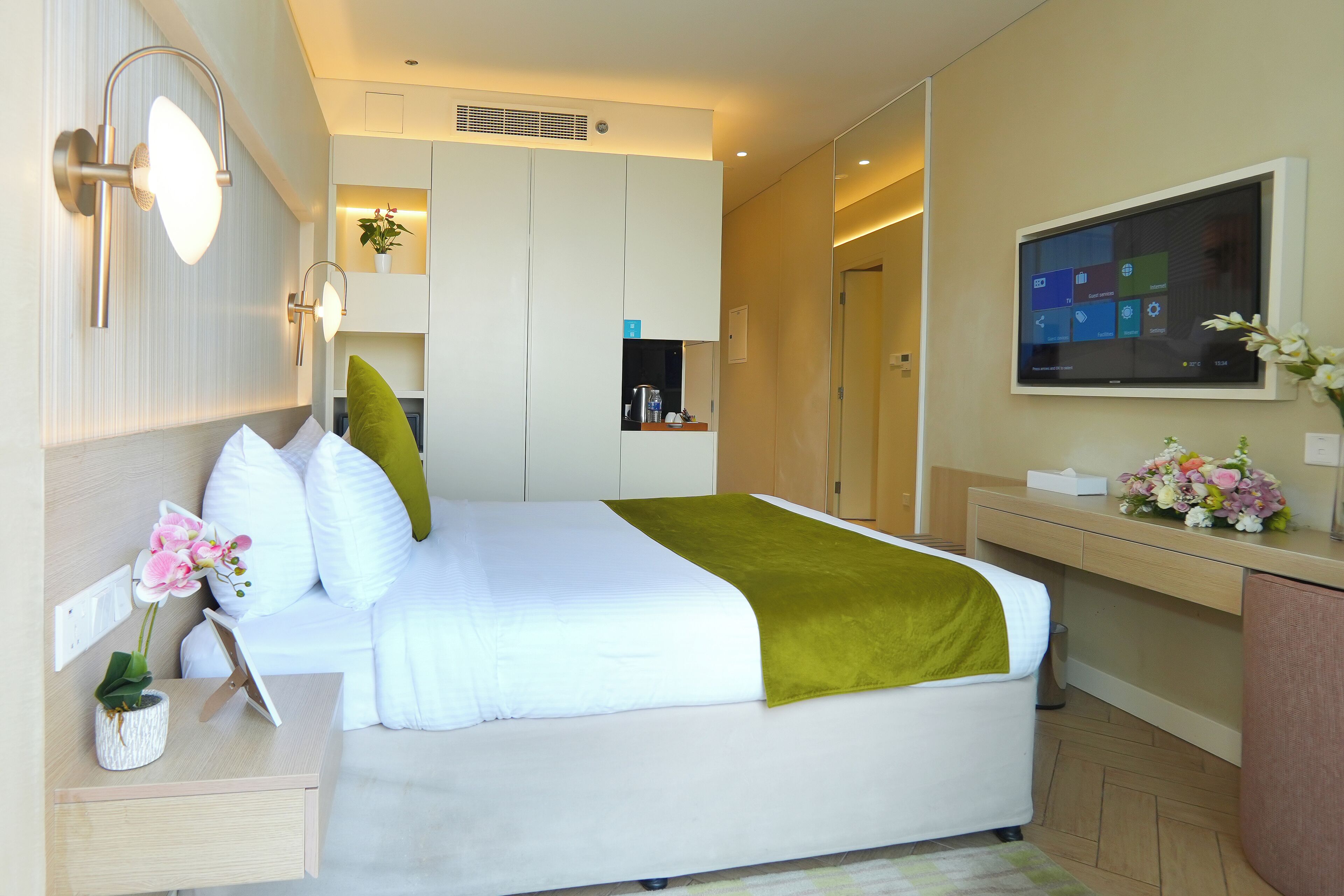 Una camera d'albergo moderna con un letto rifatto, un comodino con una lampada, un piccolo ripiano con una pianta e una televisione montata a parete.