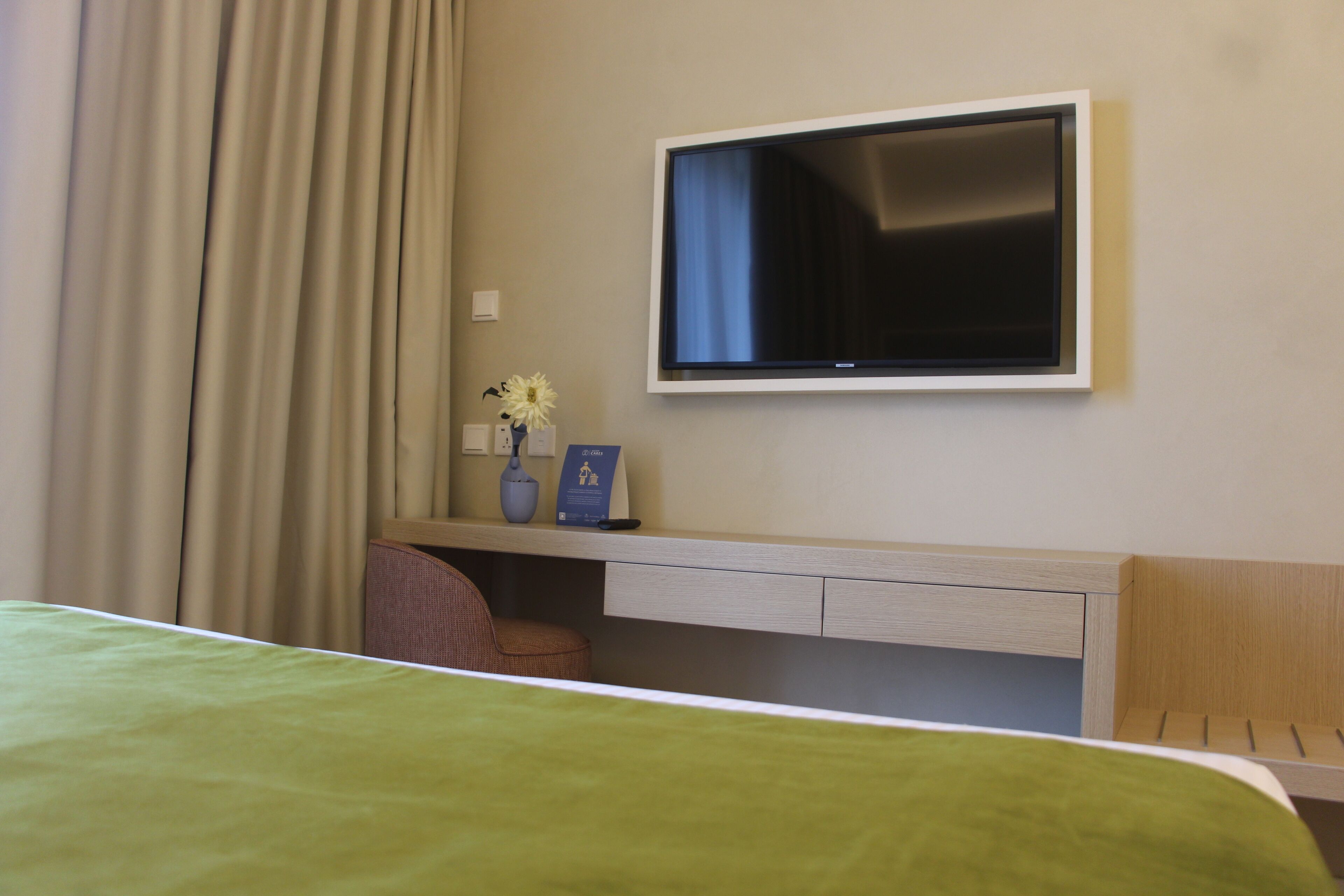 Habitación de hotel con cama, escritorio con televisor de pantalla plana, una silla y un florero con flores.