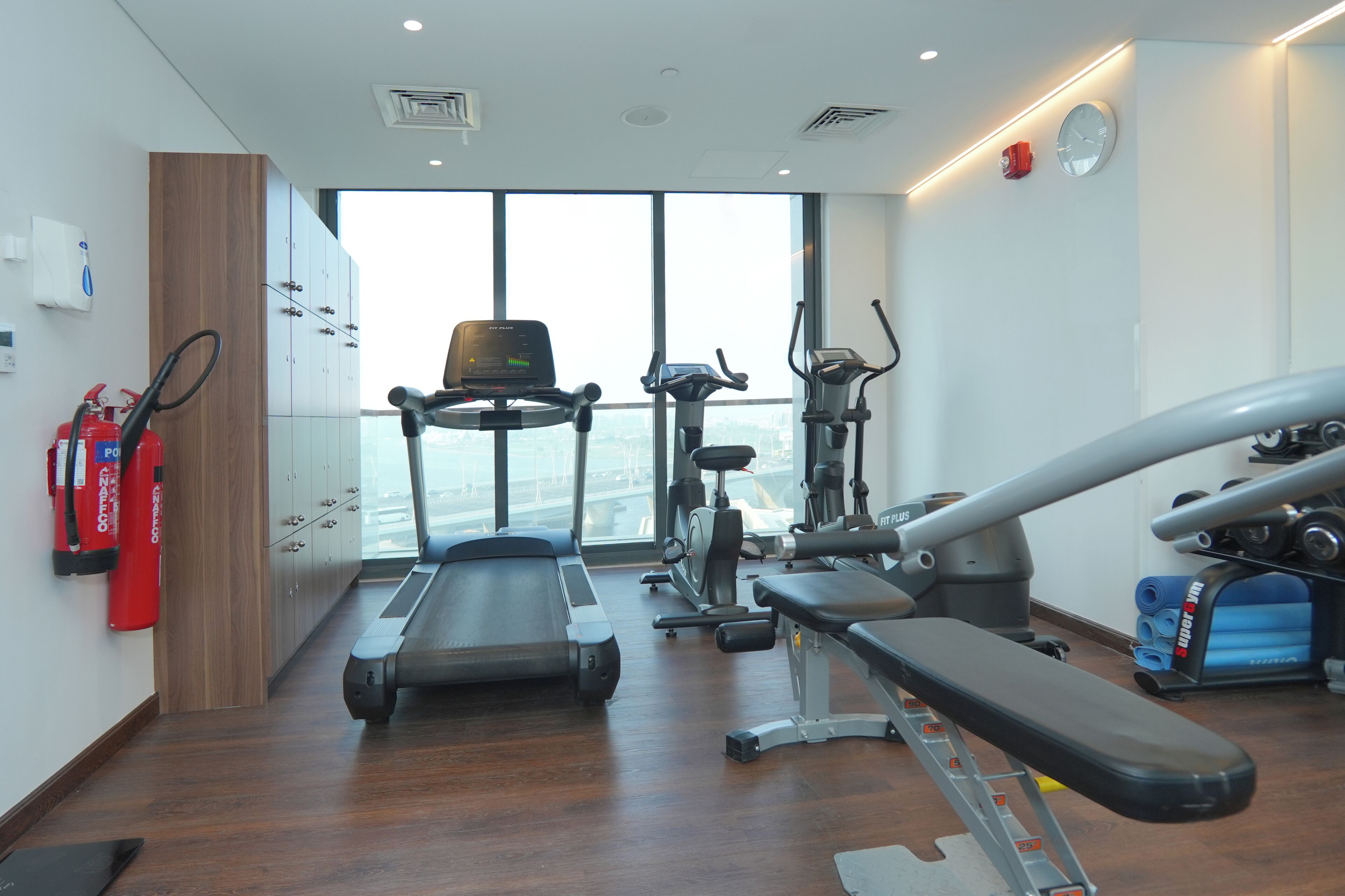 Une salle de sport équipée de machines de cardio, de bancs de musculation et d’une grande fenêtre donnant sur la ville.