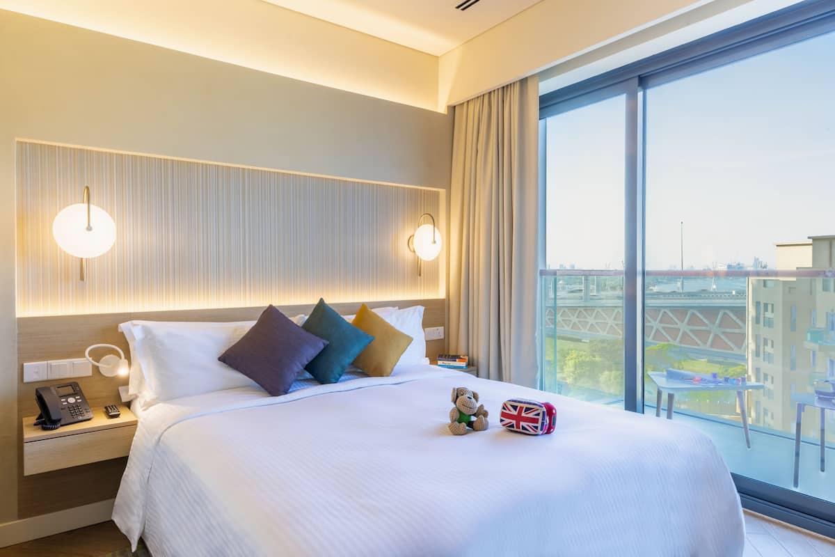 A modern hotel room with a large bed, bedside lamps, a telephone, and a view of the city through large windows.