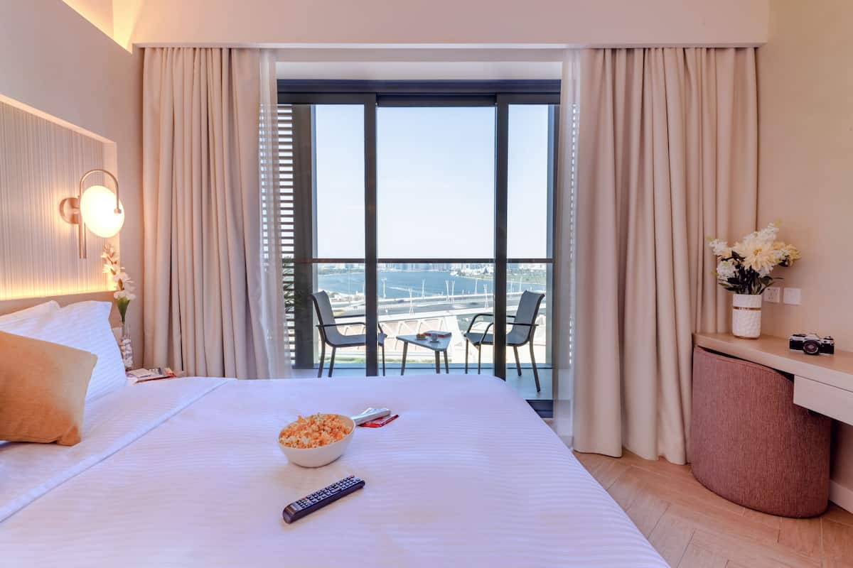 A hotel room with a large bed, a balcony with a view of the water, a desk with a camera, and a bowl of popcorn on the bed.