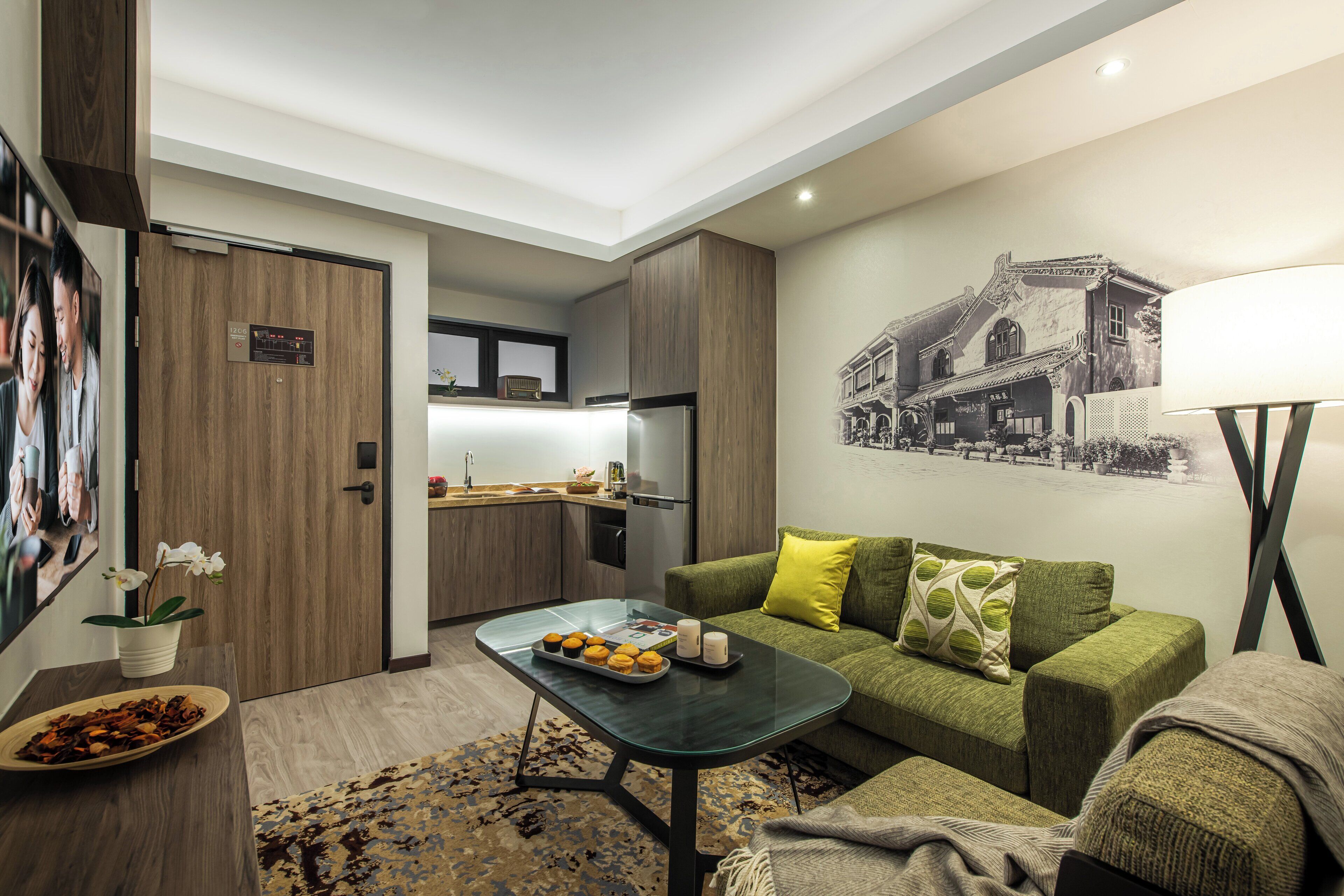 Ein modernes Wohnzimmer mit einem grünen Sofa, einem Couchtisch mit Snacks, einer sichtbaren Küche im Hintergrund und einer Wandlampe.
