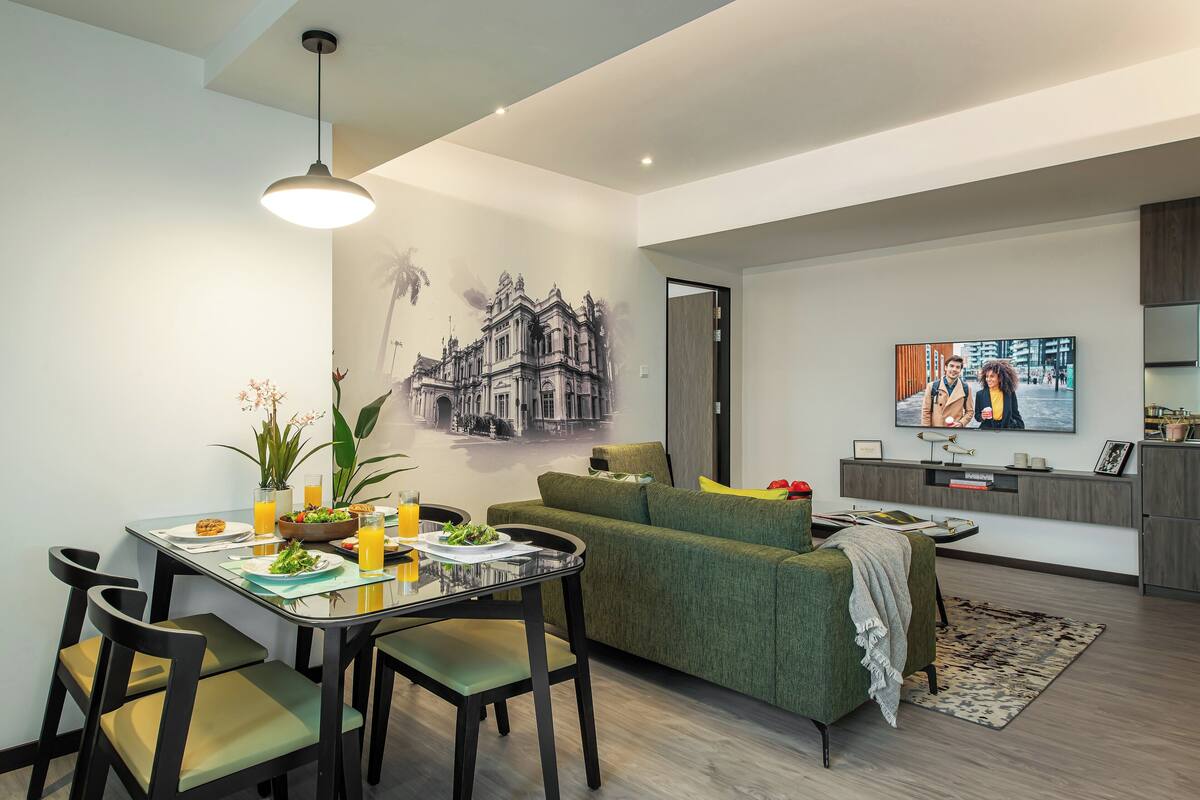A modern living room with a dining area, a green sofa, a flat-screen TV, and a kitchen area with a built-in oven.